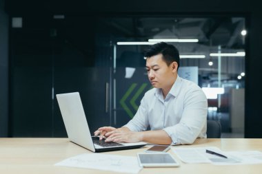 Genç yakışıklı bir adam, Asyalı bir işadamı, bir dizüstü bilgisayarla çalışıyor, daktilo ediyor. Modern bir ofiste masa başında oturmak.