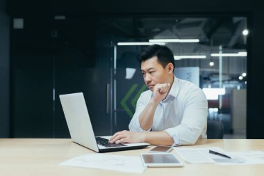 Genç yakışıklı bir adam, Asyalı bir işadamı, bir dizüstü bilgisayarla çalışıyor, daktilo ediyor. Modern bir ofiste masa başında oturmak.