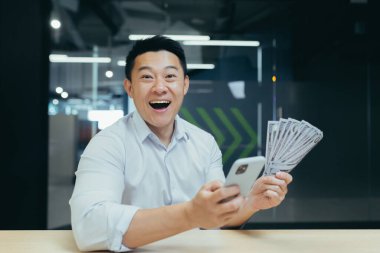Asyalı mutlu genç bir adam elinde telefon ve nakit para tutuyor. Kazandığı için mutlu. Modern bir ofiste masa başında oturmuş gülüyorum.