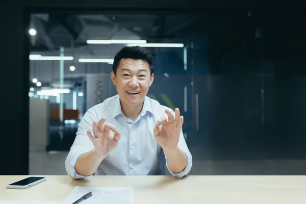 Online business training. Portrait of an active young Asian businessman. Conducts online business training on camera, sits at a table in a modern office, talks, explains, waves his hands, smiles