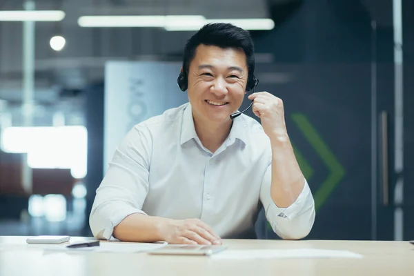 Business coach. A young handsome Asian man in headphones with a microphone conducts business training on camera, explains, tells, smiles. Sitting behind a desk in a modern office.
