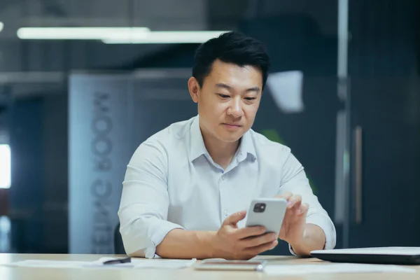 A break at work. A young Asian businessman is using the phone at his desk in a modern office. He holds it in his hands, types a message, makes a phone call.