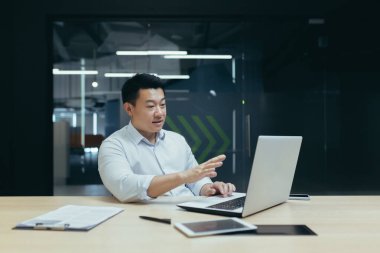 Ciddi bir genç adam, Asyalı bir işadamı, serbest çalışan, müşterileri ve ortaklarıyla bir laptoptan video görüşmesi yapıyor. Modern bir ofiste masa başında oturmak.