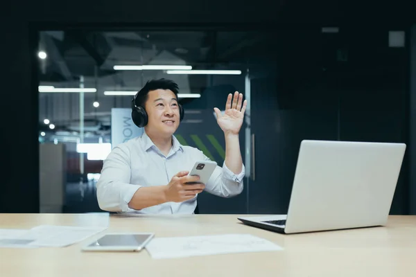 Young man Asian businessman listens to music in headphones and on the phone, took a break from work. He sits in a white shirt in a modern office at a table, dances, makes movements with his hands