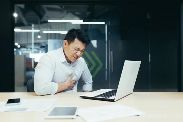 Heart attack at work. A young Asian man, businessman, office worker holds his heart, feels pain, had a stroke, heart attack at the desk in the office.