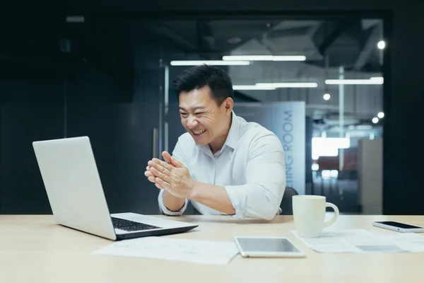 Portrait of successful Asian boss, businessman looking at laptop screen reading good news and celebrating victory and success, man at work