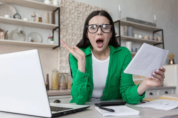 Home finances, housewife shocked looking at camera and screaming frustrated, got bad letter from bank, woman behind paper work with laptop at home in kitchen