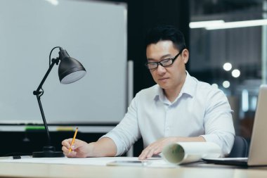 Asyalı tasarımcı modern ofiste plan üzerinde çalışıyor, erkek mimar düşünceli bir şekilde inşaat projesi üzerinde çalışıyor.