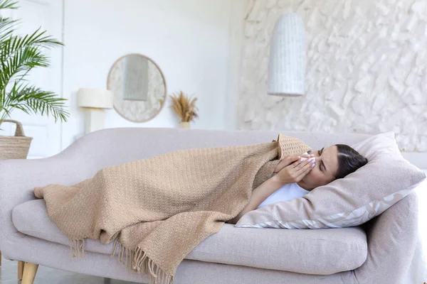 A sick woman is lying under a blanket on the sofa at home alone, has a cold, is sneezing