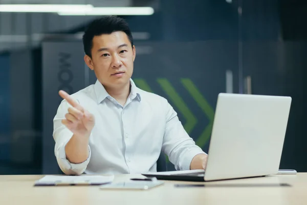 Portrait of a young Asian man, confused, shows with his hands that it is not allowed, did not understand, looks at the camera. Sitting at a desk in the office, working with a laptop online
