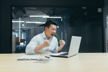 Başarılı ve mutlu Asyalı genç bir adam, iş adamı, mutlu, eliyle evet diyor. Bir anlaşma yaptık. Para kazandım. Modern bir ofiste masada bir defterle oturmak.
