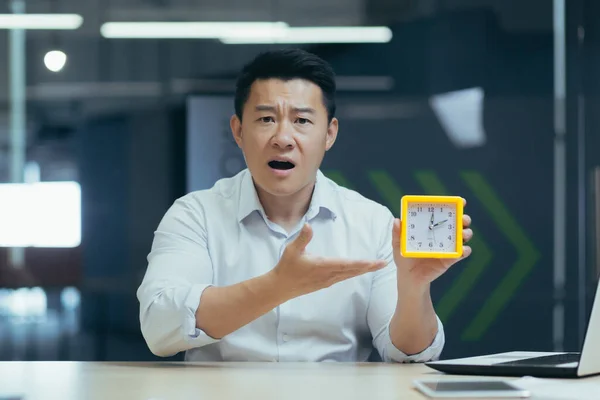 Time management. Young handsome asian businessman holding a desk clock in his hand, pointing to the clock, indicating the time, confused, late, looking at the camera