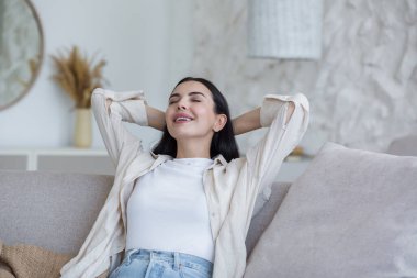 Close-up photo of a young beautiful woman relaxing at home with her eyes closed, her hands behind her head and daydreaming while sitting on the couch in the daytime