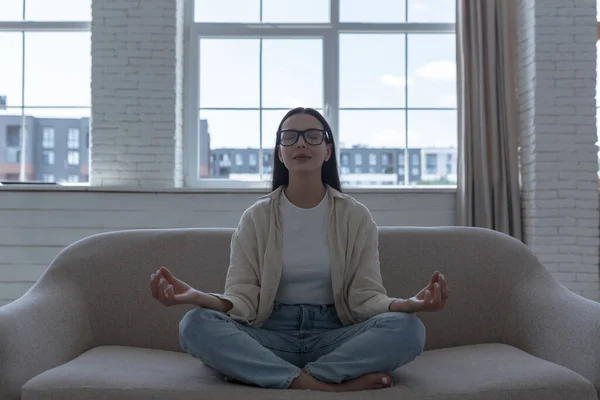 Woman alone at home meditating in front of window sitting on sofa, beautiful girl calm down and resting in lotus pose, wearing glasses with closed eyes
