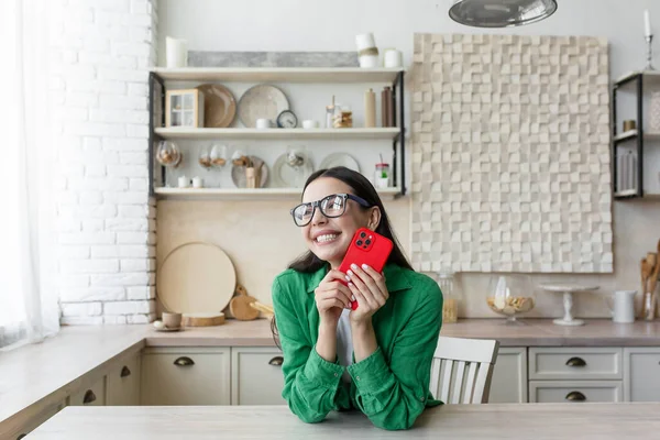 Young beautiful woman in glasses and green shirt at home got a good news, read message from red smartphone, happy and smiling in kitchen at home