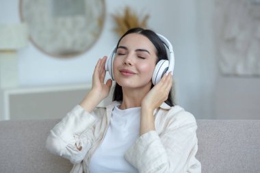 Beautiful young woman resting listening to music in white big headphones, brunette sitting on sofa in living room with closed eyes dreaming at home