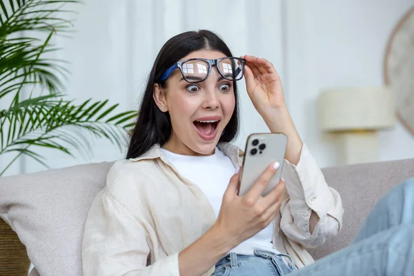 Young beautiful woman in glasses joyfully reading message from mobile phone, brunette at home sitting on sofa resting