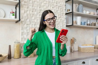 Beautiful young woman in glasses and green shirt read good news from red phone, brunette at home in kitchen shocked by good news