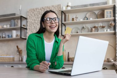Mutfakta gözlüklü güzel bir kadının portresi, bilgisayarını online alışveriş için kullanan esmer, gülümseyen ve kameraya bakan, banka kredi kartı tutan