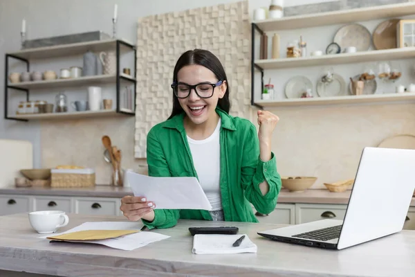 Happy woman in glasses and green shirt at home doing paper work got good result, looking at documents and happy, holding hand up gesture of success and victory