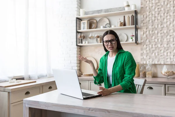 Portrait of woman at home, annoyed brunette in green shirt and glasses looking at camera and arguing, working in kitchen with laptop