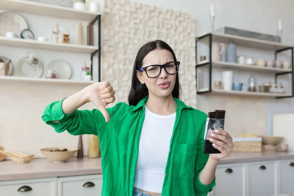 Woman at home disapproving of a bar of chocolate, looking at the camera and pointing a finger down, brunette at home in the kitchen in a green shirt and glasses