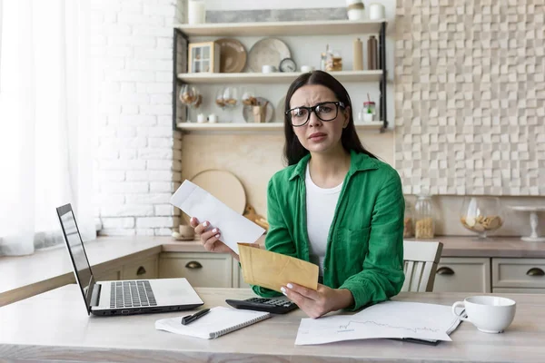 Portrait woman at home, received bad news a letter from the bank, brunette doing paperwork at home in the kitchen, counting bills and summarizing financial documents, shocked bad result looks camera