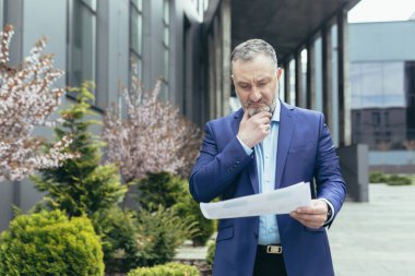 Senior and experienced gray haired boss man near office pensive reading financial report document outside office, businessman thinking