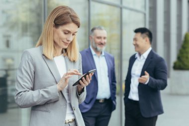 A beautiful and successful business woman is using the phone outside the office, on the photo are male colleagues talking, a successful business team is on a break
