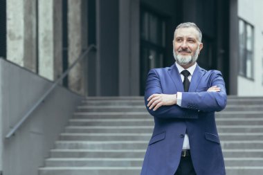 Portrait of successful director and business owner, man with crossed arms smiling outside office building, businessman investor in business suit confident
