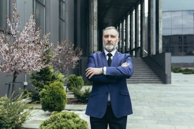 Portrait of successful thinking experienced senior businessman, gray haired male boss in business suit outside office, looking at camera with crossed arms