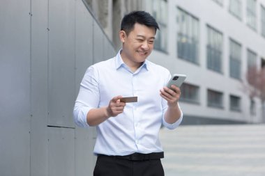 Asian businessman smiling and happy doing online shopping outside office building, man using mobile phone, holding bank credit card