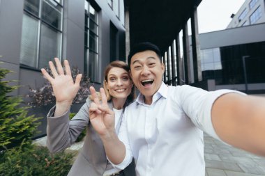 Two friends colleagues workers asian man and business woman looking at smartphone camera and waving greeting, business people talking on video call smiling and happy