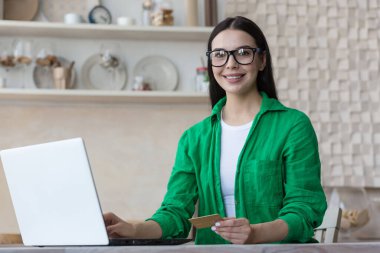 Online shopping. Portrait of a beautiful woman in glasses and a green shirt sits at home with a laptop, holds a credit card in her hands. Makes purchases on the Internet, orders delivery, pays online.