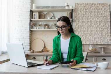 Home accounting.A young woman counts her income and expenses.checks the household budget. In glasses and a green shirt, sits at home with a laptop,a calculator and writes in a notebook,calculates.