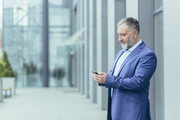 Serious and confident gray-haired man standing outside office building, businessman senior banker in business suit holding phone, successful investor using smartphone