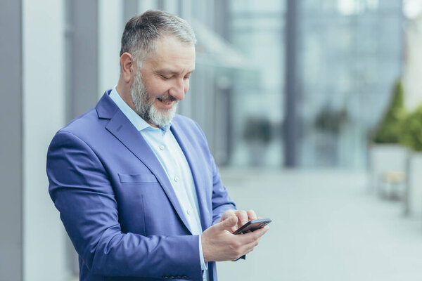 Successful senior and experienced gray-haired man outside office building reading message on phone, businessman banker in business clothes smiling and happy, investor on day break