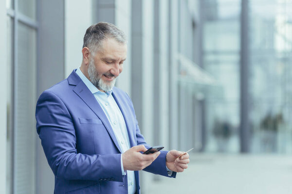 Senior gray-haired businessman outside office building, man in business suit uses mobile phone smartphone to buy in online store and order service, holds bank card in hands