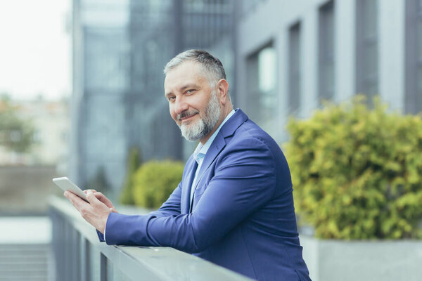 Portrait of successful senior gray-haired businessman boss, man outside office using tablet computer smiling and looking at camera, investor in business suit, happy banker