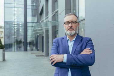 Portrait of serious senior gray-haired investor, man with crossed arms outside office looking at camera, businessman in glasses and business suit