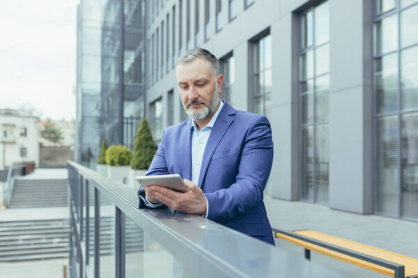 Successful and serious pensive senior gray haired man outside office building using tablet computer, businessman investor reading news from tablet