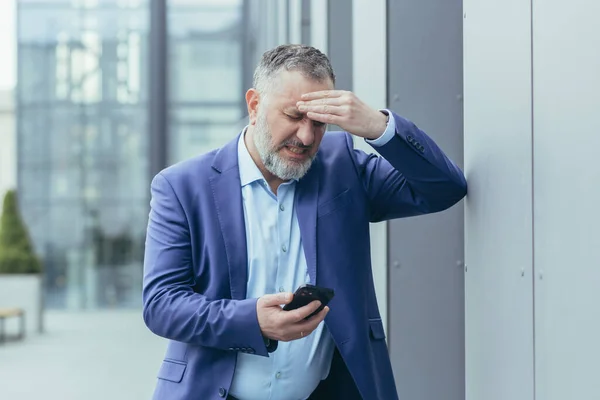 Senior gray-haired businessman sick outside office building, man holding hand to head, having severe headache, boss trying to call doctor on phone and call ambulance