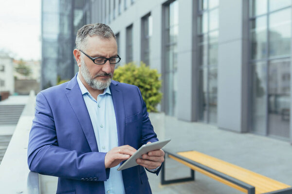 Concentrated and serious senior gray-haired banker using tablet computer outside office building, businessman reading news and thinking, man in glasses and business suit typing message
