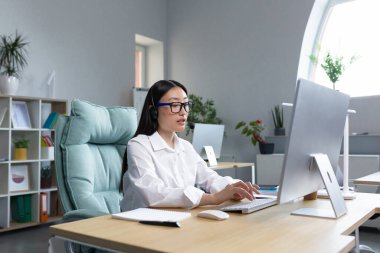 Çevrimiçi mesafe öğrenimi. Kulaklıklı, gözlüklü, genç ve güzel Asyalı bayan öğretmen bilgisayarın başında oturmuş internette ders veriyor..