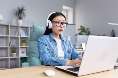 Online training. Young beautiful Asian girl student in glasses studying at a laptop in headphones. Learns remotely, sits at a desk in a modern office.
