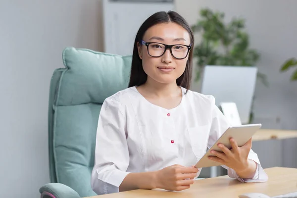 Online admission to the hospital. A young beautiful Asian woman doctor records a patients medical history online in a tablet. Typing in a tablet. Sitting at the table in the winding office.