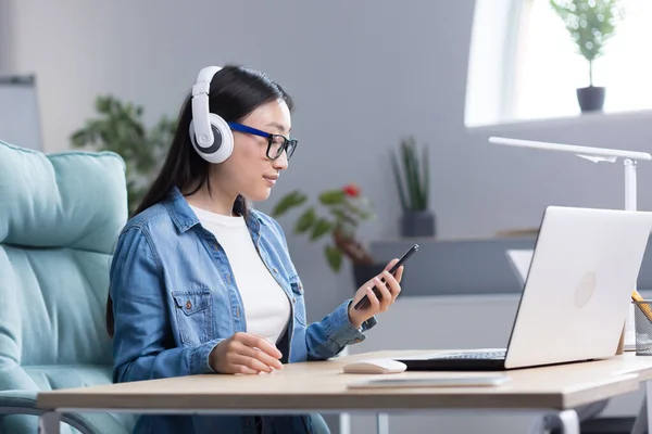 Video chat with friends. Young beautiful Asian woman wearing headphones, holding a phone in her hand, talking on a video call with friends. Sitting at a table with a computer in a office. Smiling.