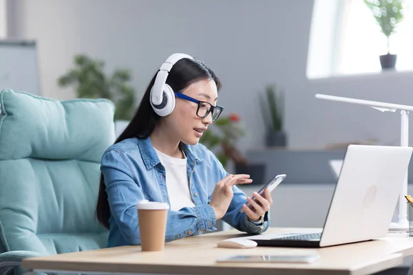 Online training. A young beautiful Asian student girl in glasses is studying at a laptop in headphones, holding a phone in her hands. Learns remotely, sits at a desk in a modern office.
