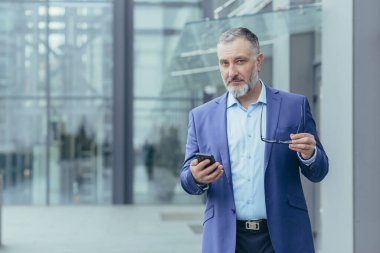 Portrait of senior gray-haired financier, man outside office building holding phone and looking at camera, businessman walking
