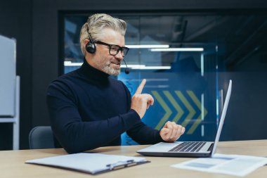 Senior and experienced gray-haired boss working in modern office, man with headset talking on video call using laptop while sitting at table, businessman in online meeting talking colleagues remotely